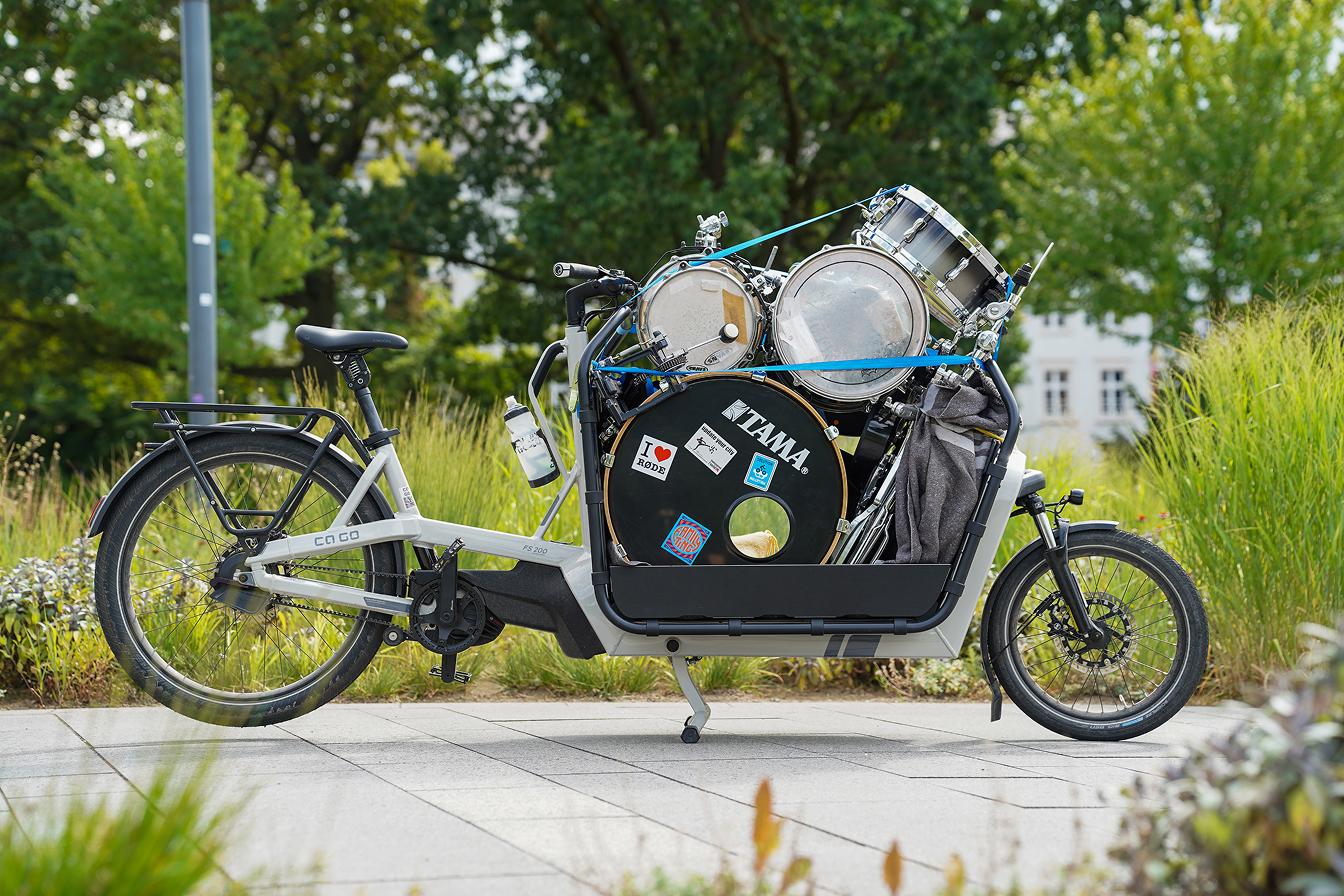 Ein mit einem Schlagzeug beladenes und mit Gurten gesichertes Lastenfahrrad, das auf einem gepflasterten Weg inmitten von Grünflächen abgestellt ist.